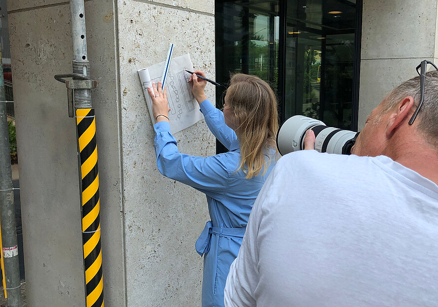 Zwei Personen stehen draußen. Eine Person zeichnet etwas auf eine Papier an der Wand. Eine Person hält eine Kamera.