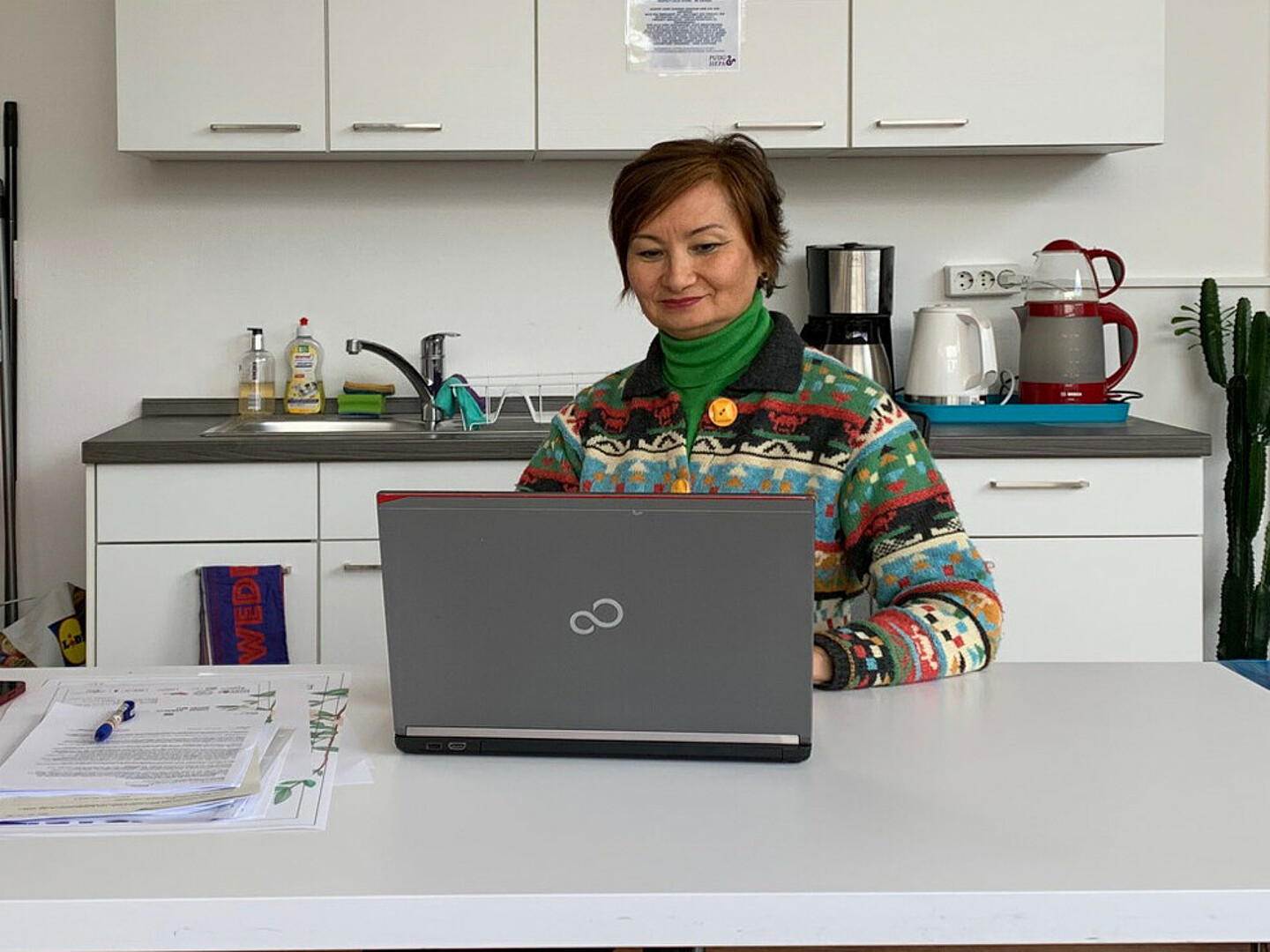 Alma Evert sitzt am Schreibtisch und schaut auf ihren Laptop, neben ihr ein Stapel Papiere.