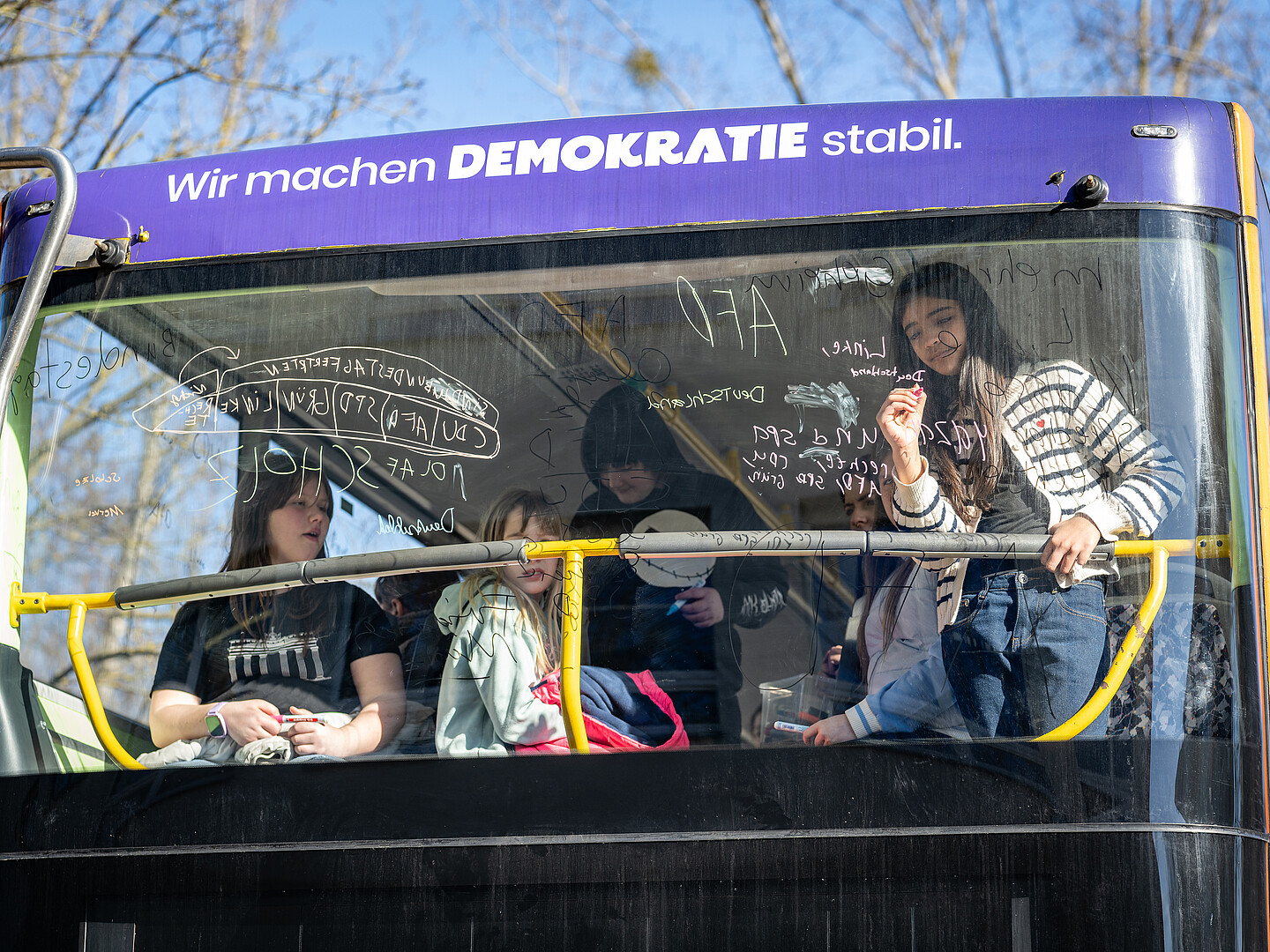 Mehrere Kinder stehen und sitzen hinter der Frontscheibe des Demokratie-Mobils und schreiben etwas mit Fensterstiften an die Scheibe.