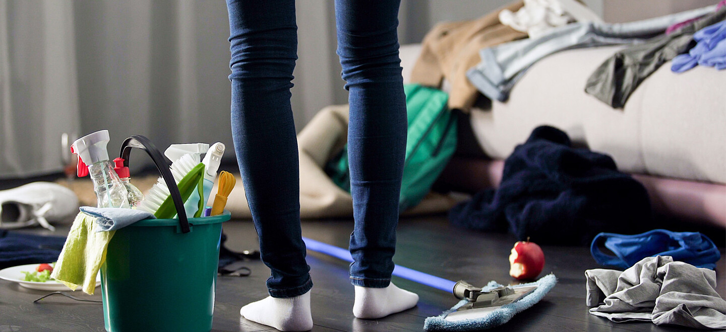 Foto aufgenommen von oberhalb des Knies einer Person nach unten. Unter dem Boden befinden sich Reinigungswerkzeuge. Viele Klamotten werden im Haus herumgeworfen.