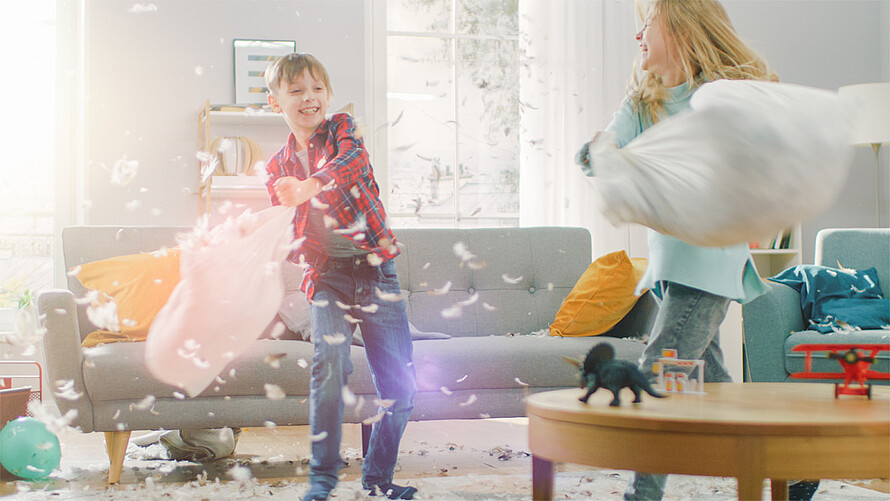 Zwei lachende Kinder machen im Wohnzimmer eine Kissenschlacht.