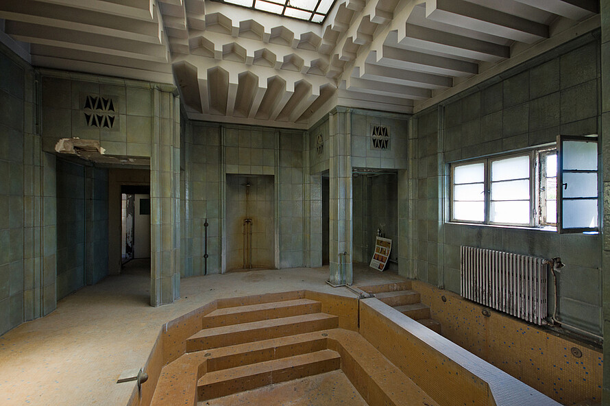 Eine alte Schwimmhalle mit orangefarbenen Fliesen und die Treppen zum Schwimmbecken sind zu sehen. Von der Decke und der Wand gelangt Licht durch die Fenster in den Raum. 