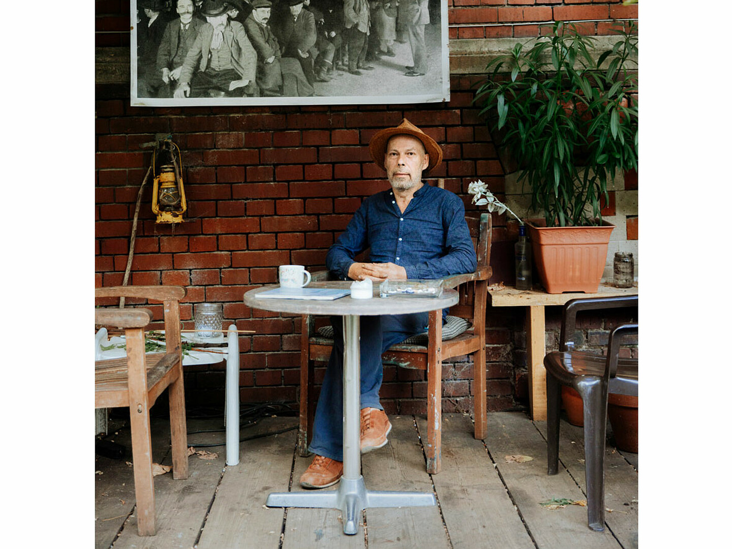 Frontaufnahmen von dem Künstler Thomas Hendriksson. Er sitzt an einem Tisch, darauf liegen verschiedene Sachen. Hinter ihm hängt ein schwarz-weißes Bild