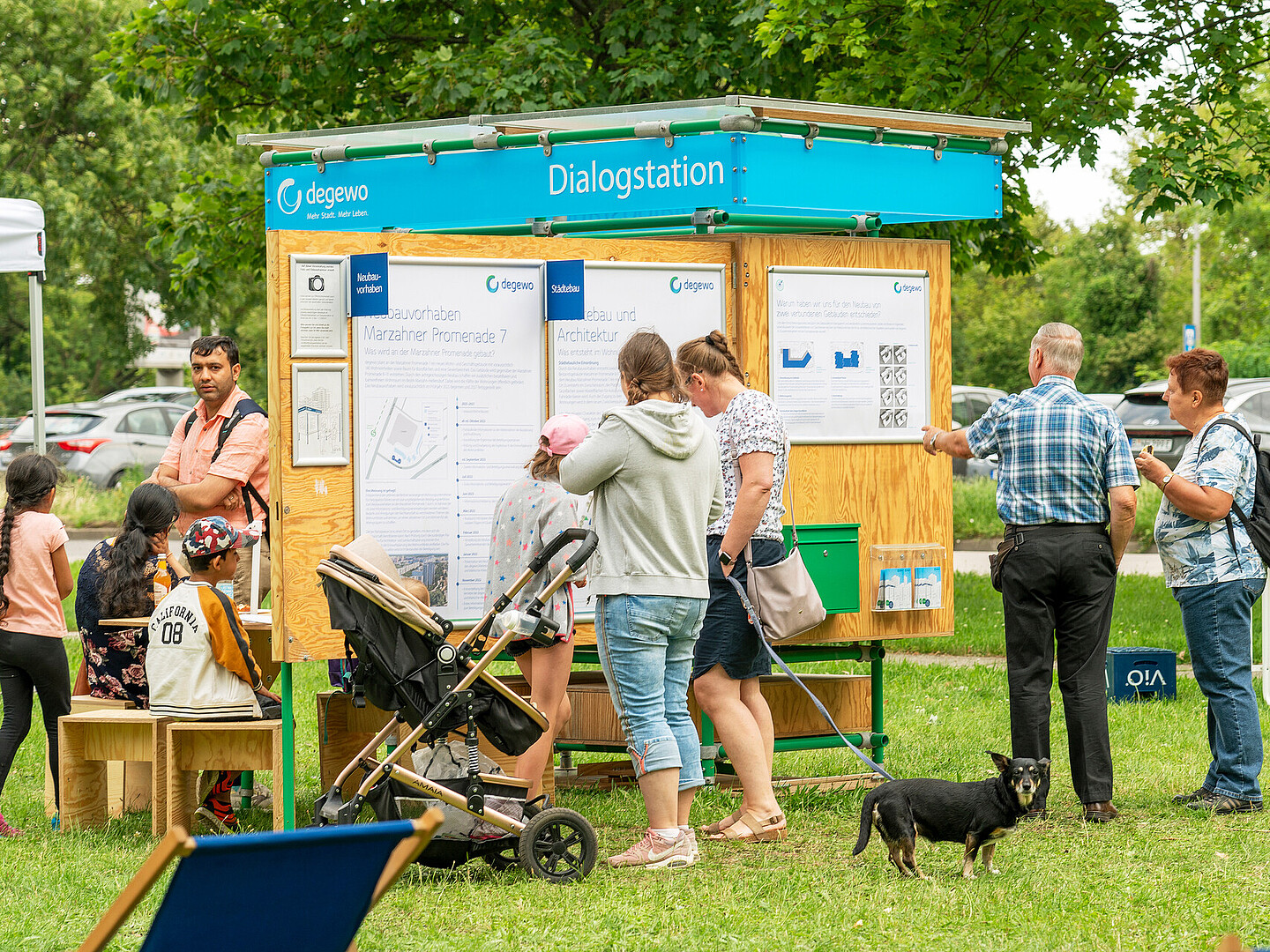 Die degewo-Dialogstation steht auf einer grünen Wiese. An den aufgeklappten Seiten hängen Plakate. Davor stehen Personen und schauen sich die Plakate an. An der hinteren Seite der Dialogstation sitzen weitere Personen an einem kleinen Tisch.
