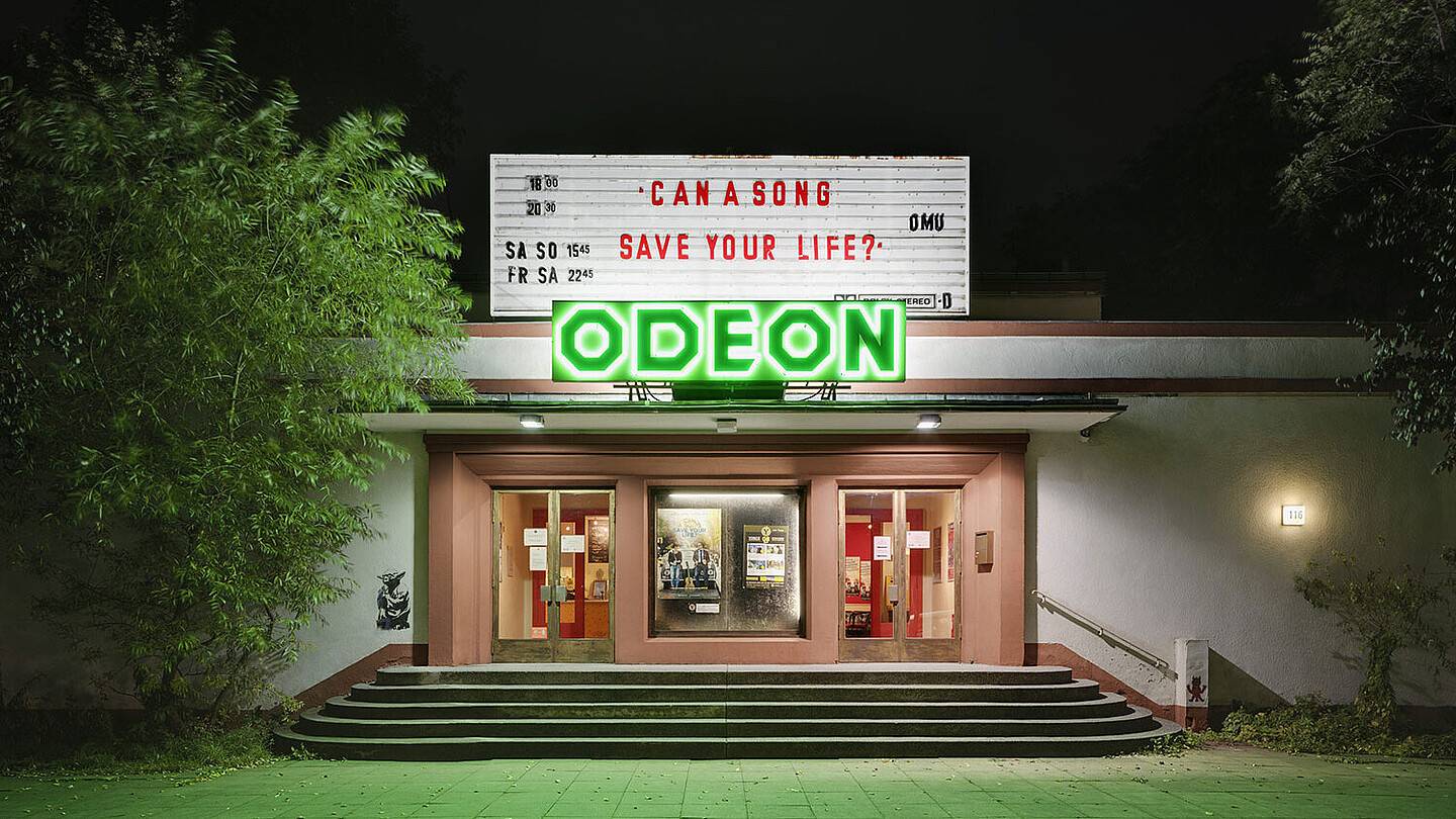 Das Odeon von außen, ein Flachbau: Das mit grüner Leuchtschrift gezierte, von der Straße zurückgesetzte Kino erreicht man über wenige Treppenstufen.