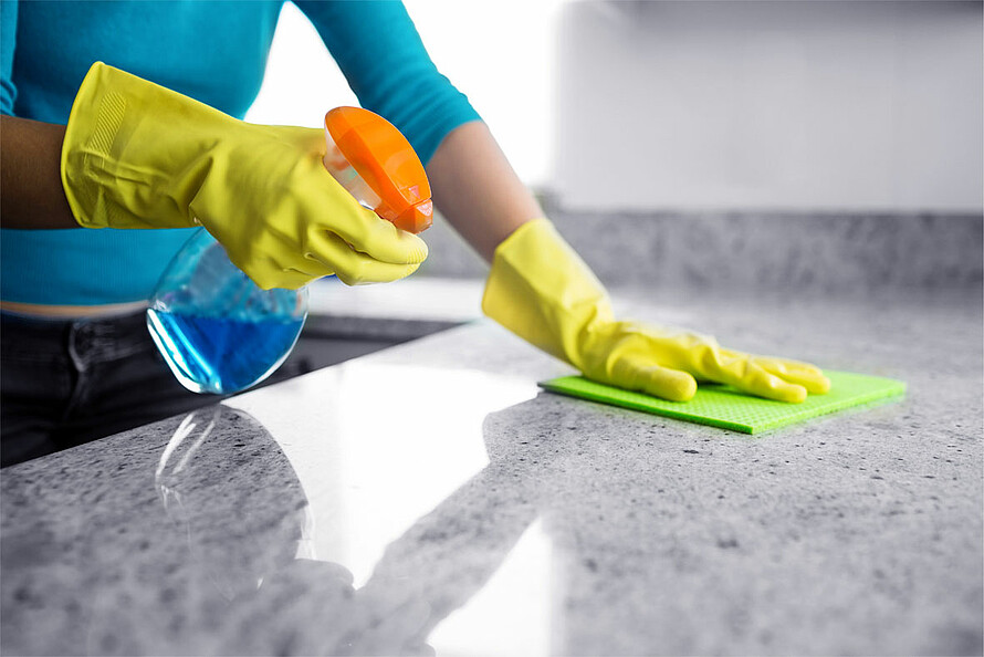 Das Foto zeigt eine Person mit gelben Gummihandschuhen, die in der rechten Hand eine Sprühflasche mit Reinigungsmittel hält und in der linken Hand einen Putzlappen.