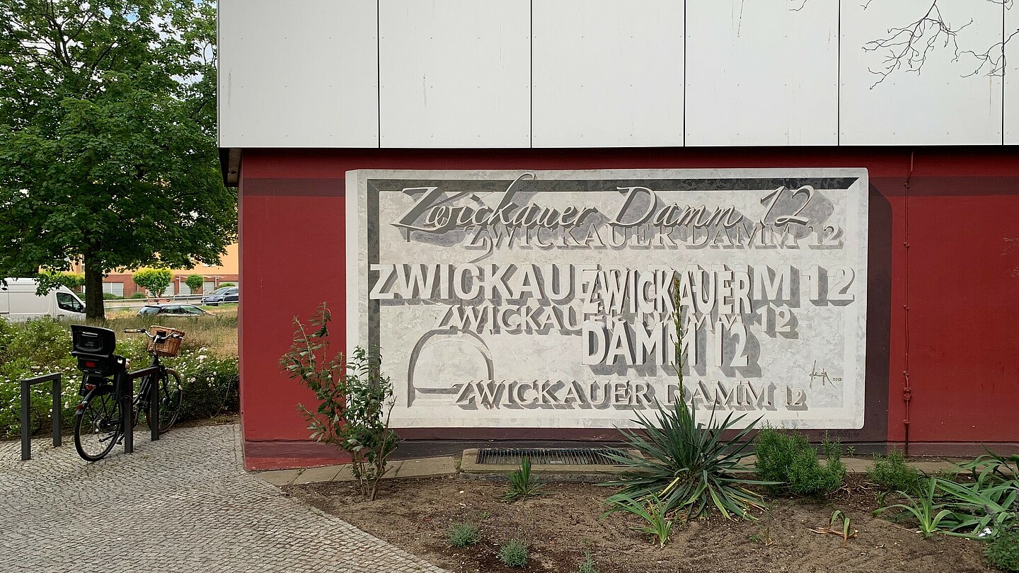 Ein rechteckiges, grau-weißes Wandgemälde auf dunkelrotem Untergrund, in dem mehrfach in verschiedenen Schriftarten „Zwickauer Damm 12“ geschrieben wurde.