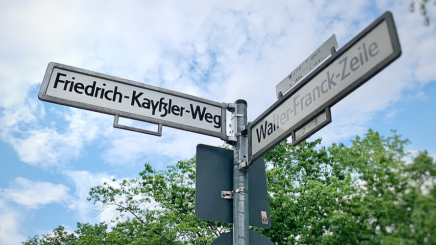 Ein Straßenschild mit der Aufschrift „Friedrich-Kayßler-Weg“. Daneben ein Straßenschild mit der Aufschrift „Walter-Frank-Zeile“. Im Hintergrund blauer Himmel, ein paar Wolken und Baumkronen.