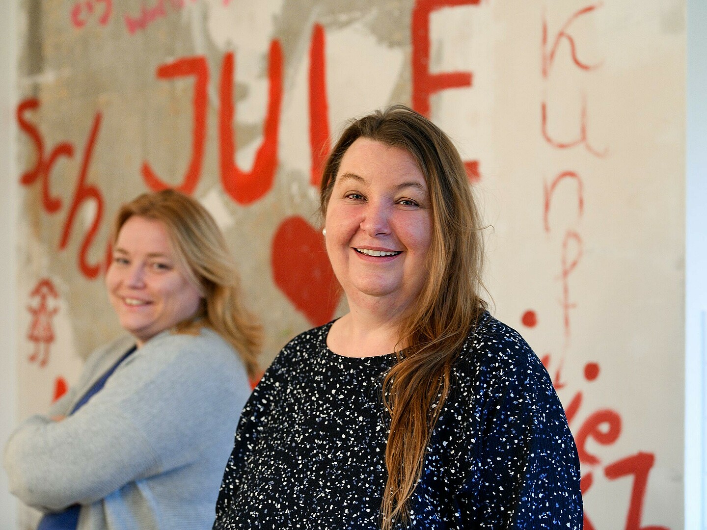 Marina Bikádi vor einer bunten Wand, auf der unter anderem „JuLe“ (Abkürzung für „Junges Leben“) steht. Im Hintergrund leicht unscharf eine weitere Person.