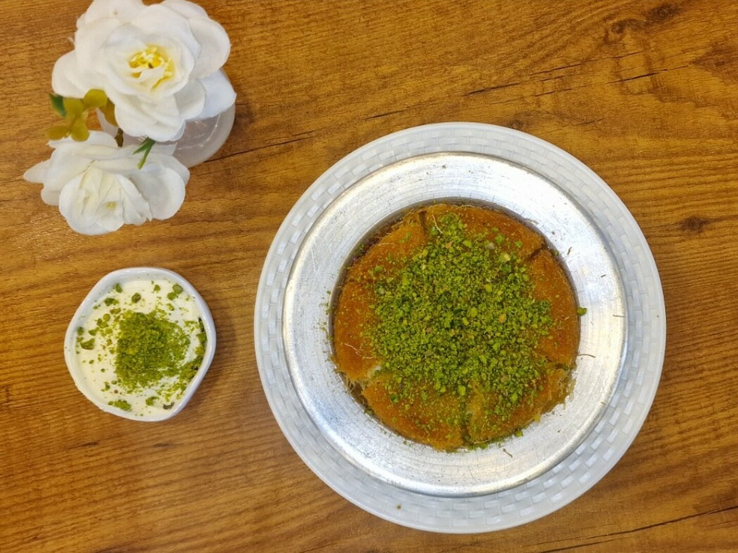 Ein Holztisch, auf dem eine Vase mit weißen Blumen und ein Teller mit Künefe stehen.