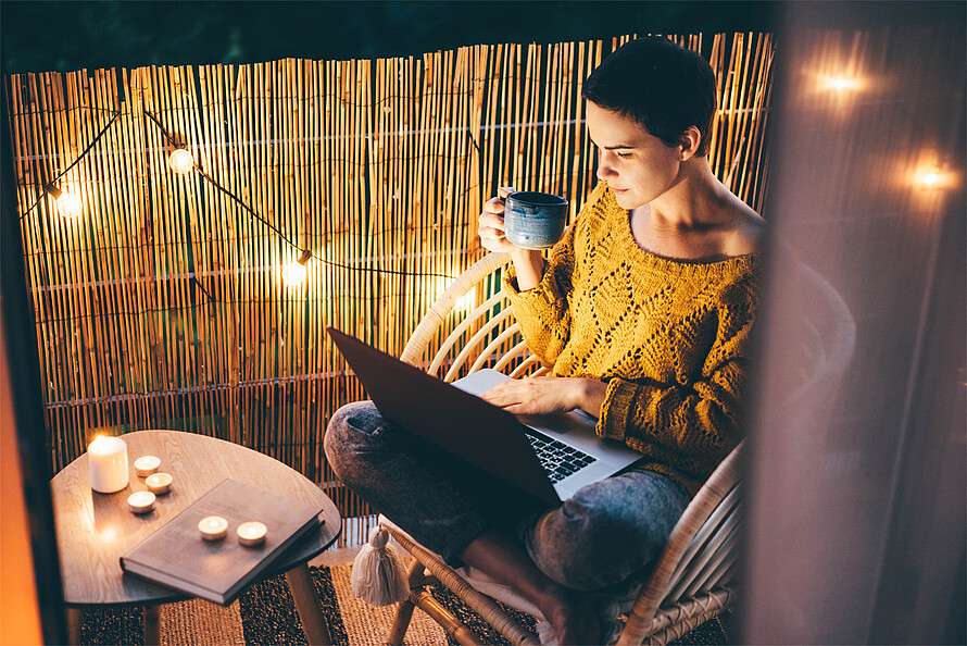 Eine junge Frau sitzt auf dem Balkon, trinkt aus einer Tasse und arbeitet an ihrem Laptop. Lichtquellen sind Kerzen und eine Lichterkette