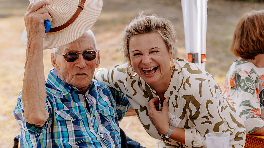 Ein älterer Herr und eine Mitarbeiterin von SOPHIA lachen in die Kamera.