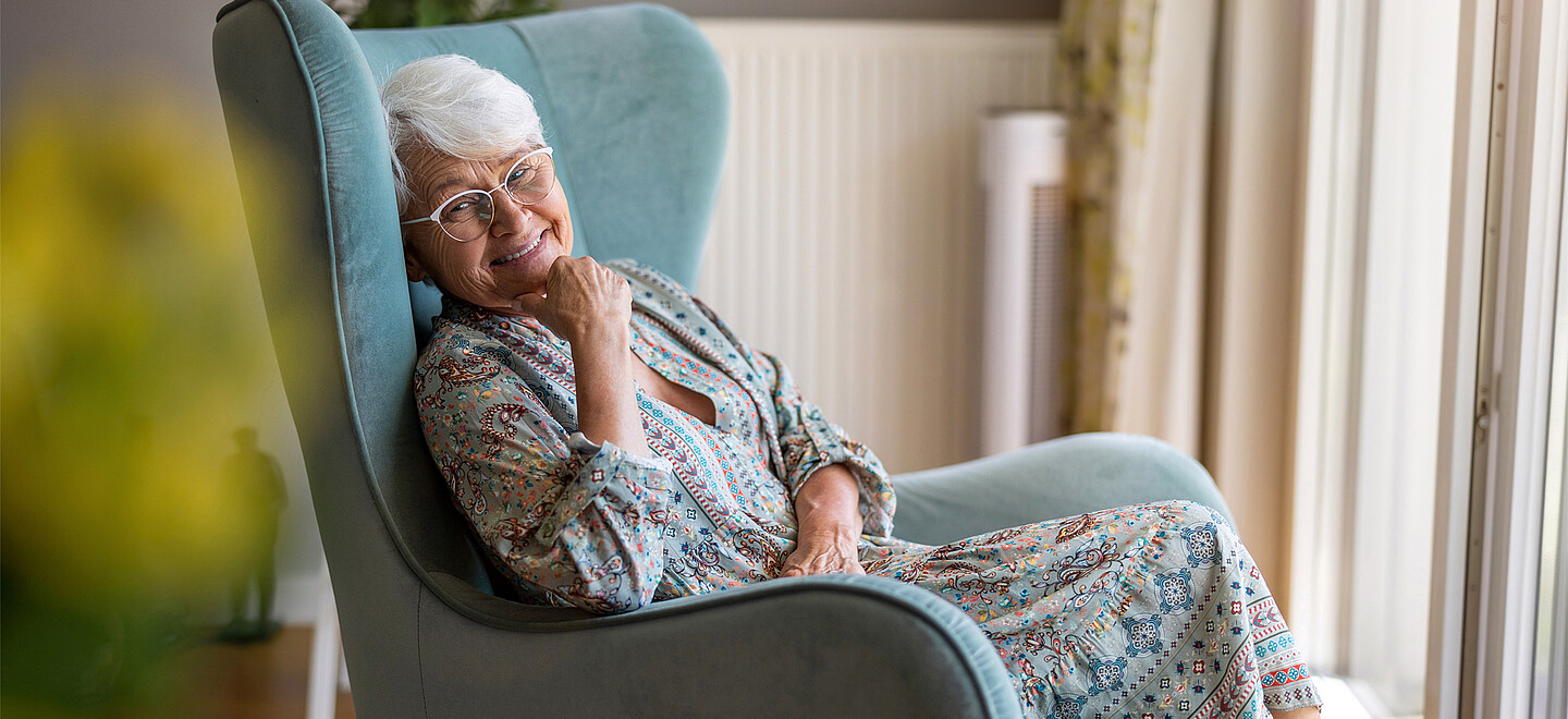Eine ältere Frau sitzt auf einem Stuhl vor dem Fenster und lächelt.