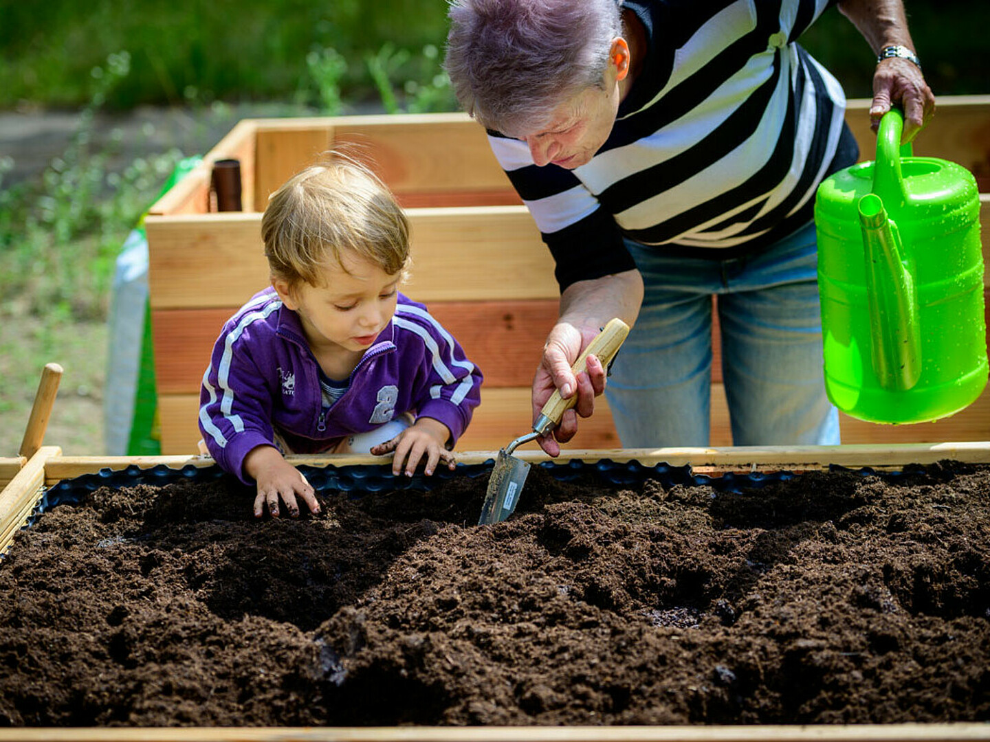 Ein Mann hält eine Gießkanne, eine Schaufel und zeigt einem Jungen, wie man ein Loch macht, um Samen darin zu pflanzen.