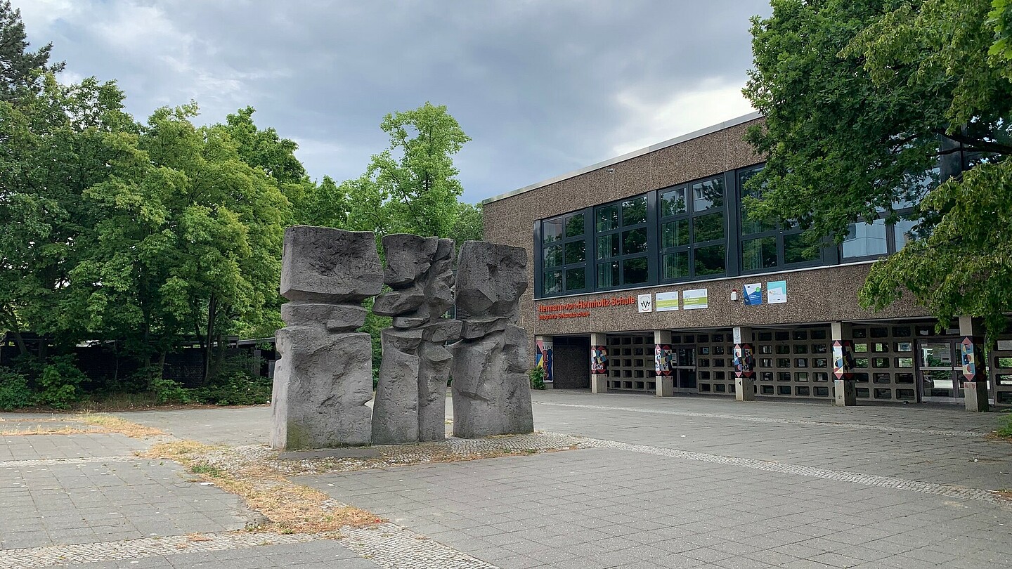 Eine Steinmauer die durch unregelmäßig geformte Löcher im Material den Eindruck erweckt, zerbrochen zu sein. Das Objekt befindet sich im Eingangsbereich eines Schulgebäudes.