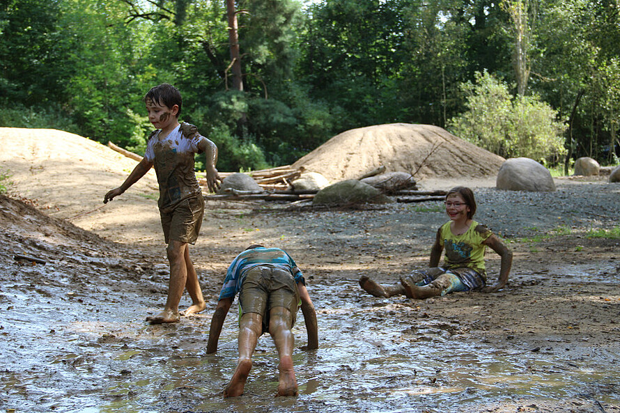 Eine Gruppe Kinder spielt im Wald. Alle drei sind mit braunem Schlamm vom Boden überdeckt.