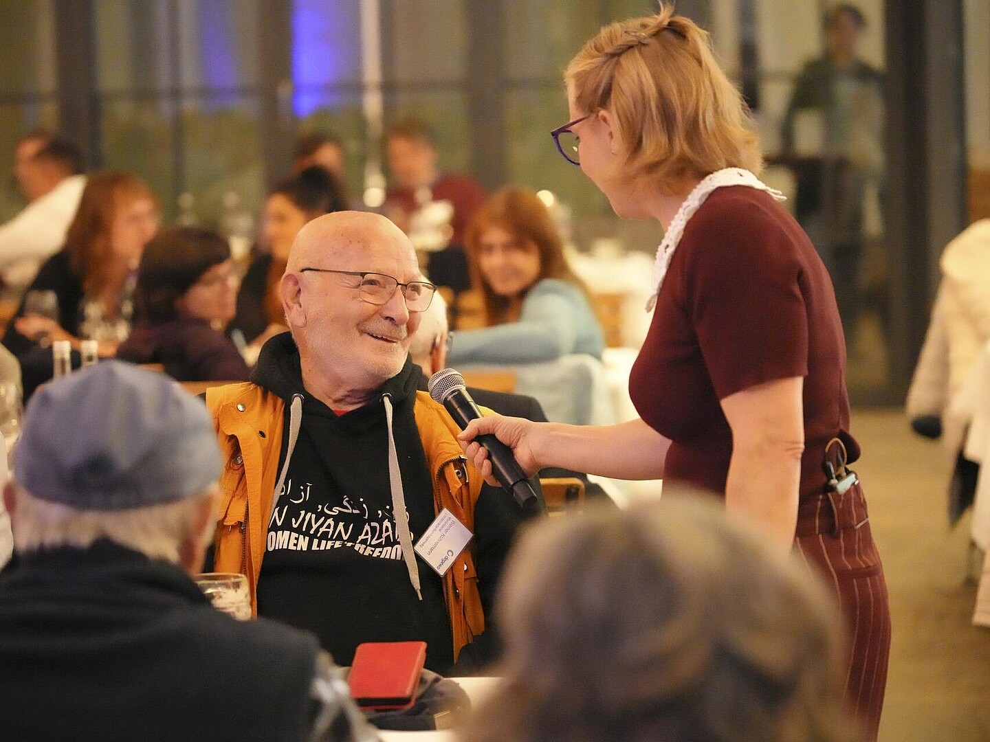 Ein Gast wird von einem Mitglied des Improvisationstheaters interviewt. Er sitzt am Tisch und lacht die Moderatorin an.