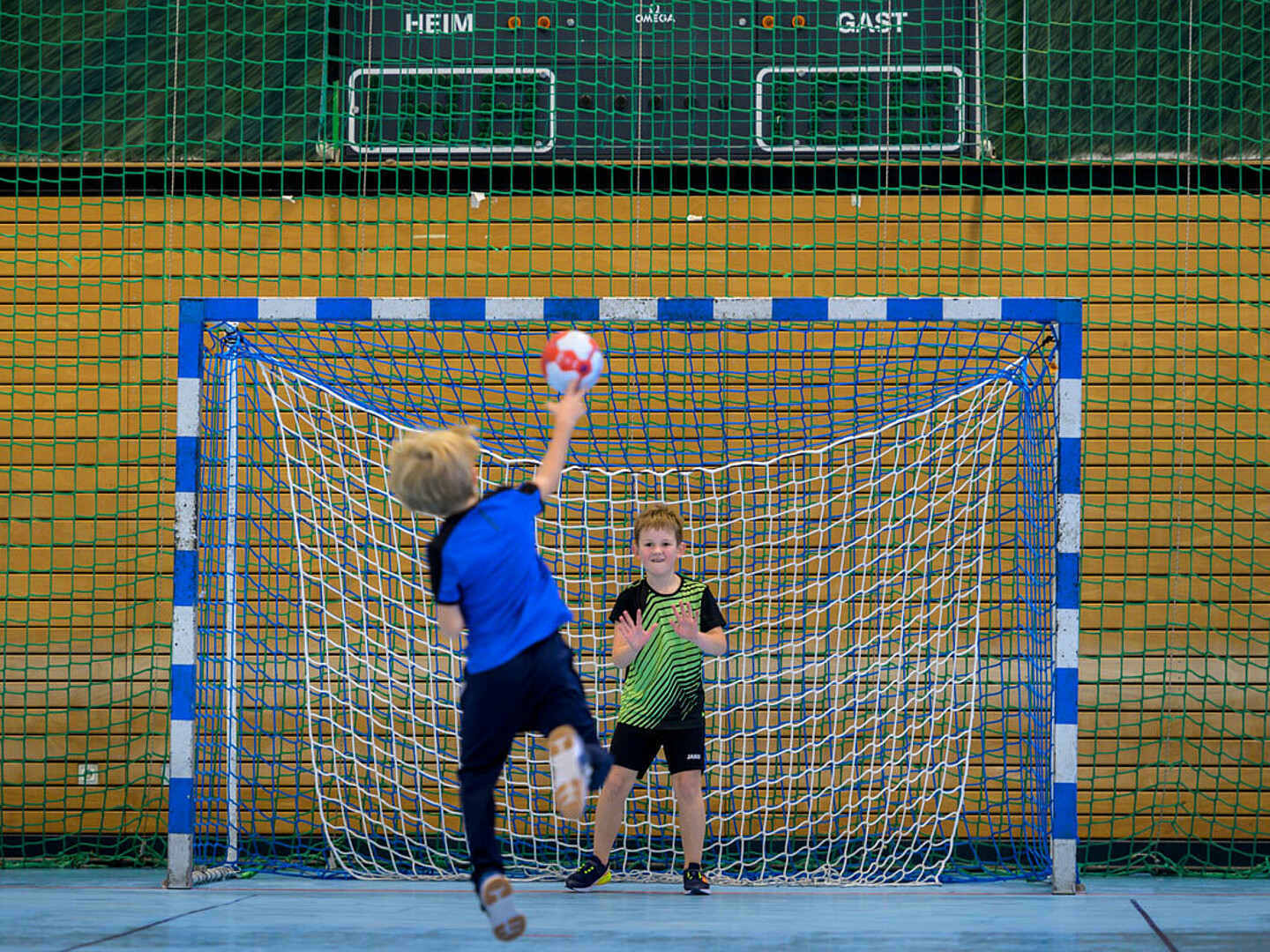 Ein Junge in Blau wirft den Ball auf das Tor und der Torwartjunge hebt seine Hand, um den Ball zu blockieren.