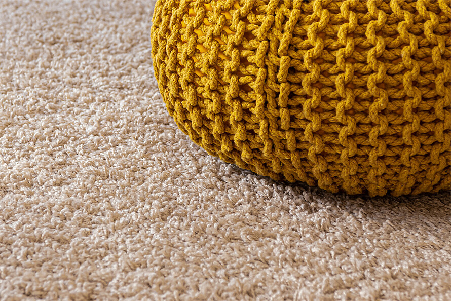 Ein cremefarbener, hochfloriger Teppich, auf dem ein senfgelbes Sitzkissen liegt.