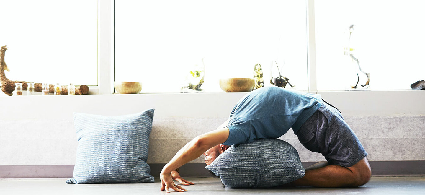 Ein Mann praktiziert Yoga in einer Backbend-Pose. er hat ein graues Kissen, um auf seinem Rücken zu ruhen. Hinter ihm befindet sich ein Fensterbrett, das mit einer Reihe von Schalen und Pflanzen geschmückt ist. Auf dem Boden liegt auch ein Kissen in der gleichen Farbe.