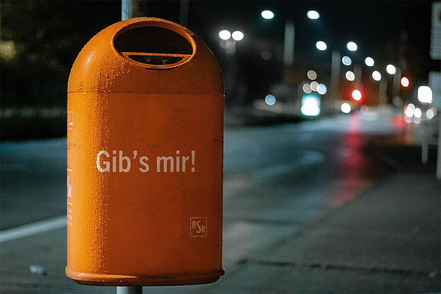 Das Foto zeigt eine Straßenszene bei Nacht, im verschwommenen Hintergrund leuchten Laternen. Auf einem orangenen Mülleimer der Berliner Straßenreinigung steht „Gib’s mir!“