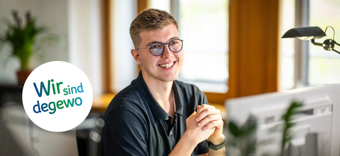 Ein junger Mann mit Brille sitzt am Schreibtisch und stützt seine Arme auf den Tisch. Er lächelt freundlich.