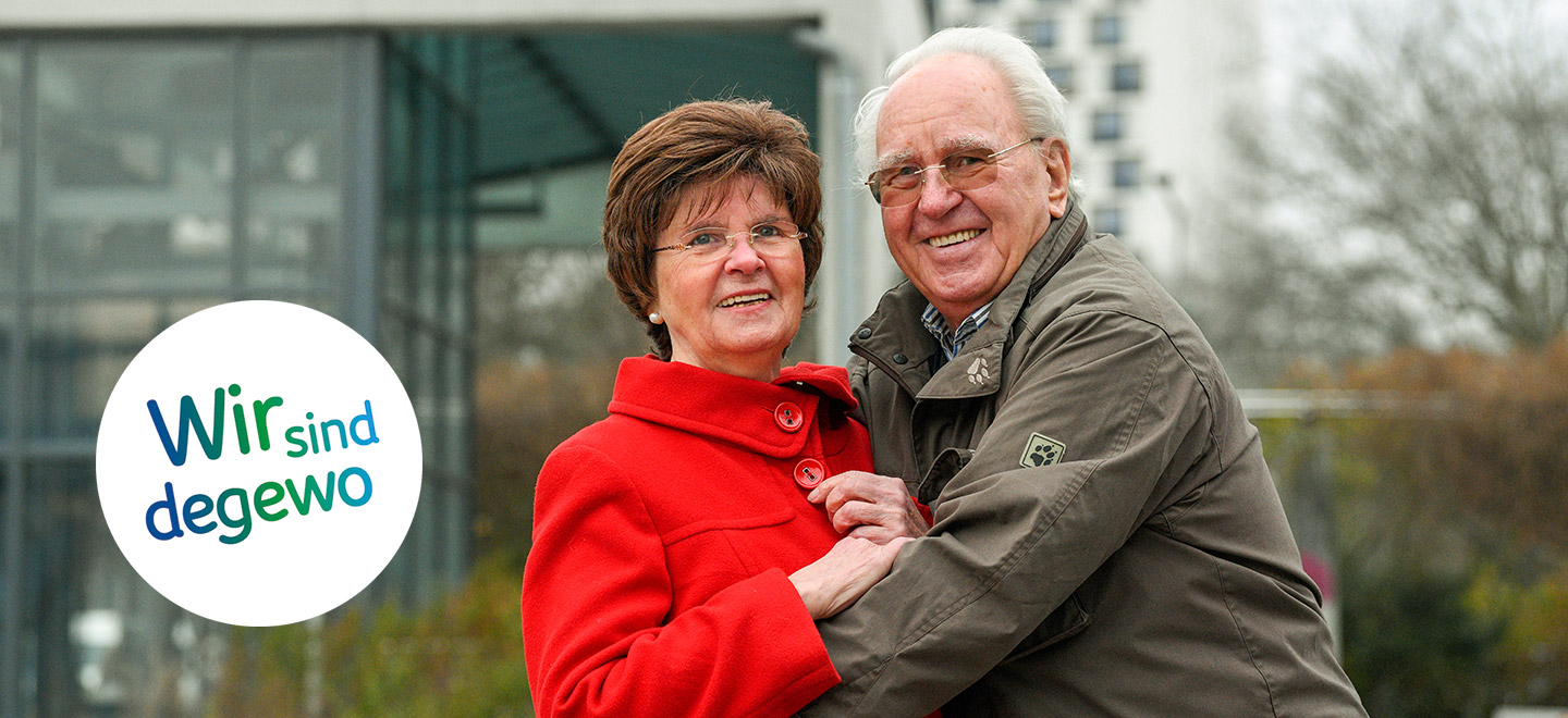 Bärbel und Udo Schulte in ihrer Nachbarschaft in Gropiusstadt. Udo Schulte trägt eine ockerfarbene Jacke und hat die Arme um seine Frau im roten Mantel gelegt. Beide lächeln in die Kamera.