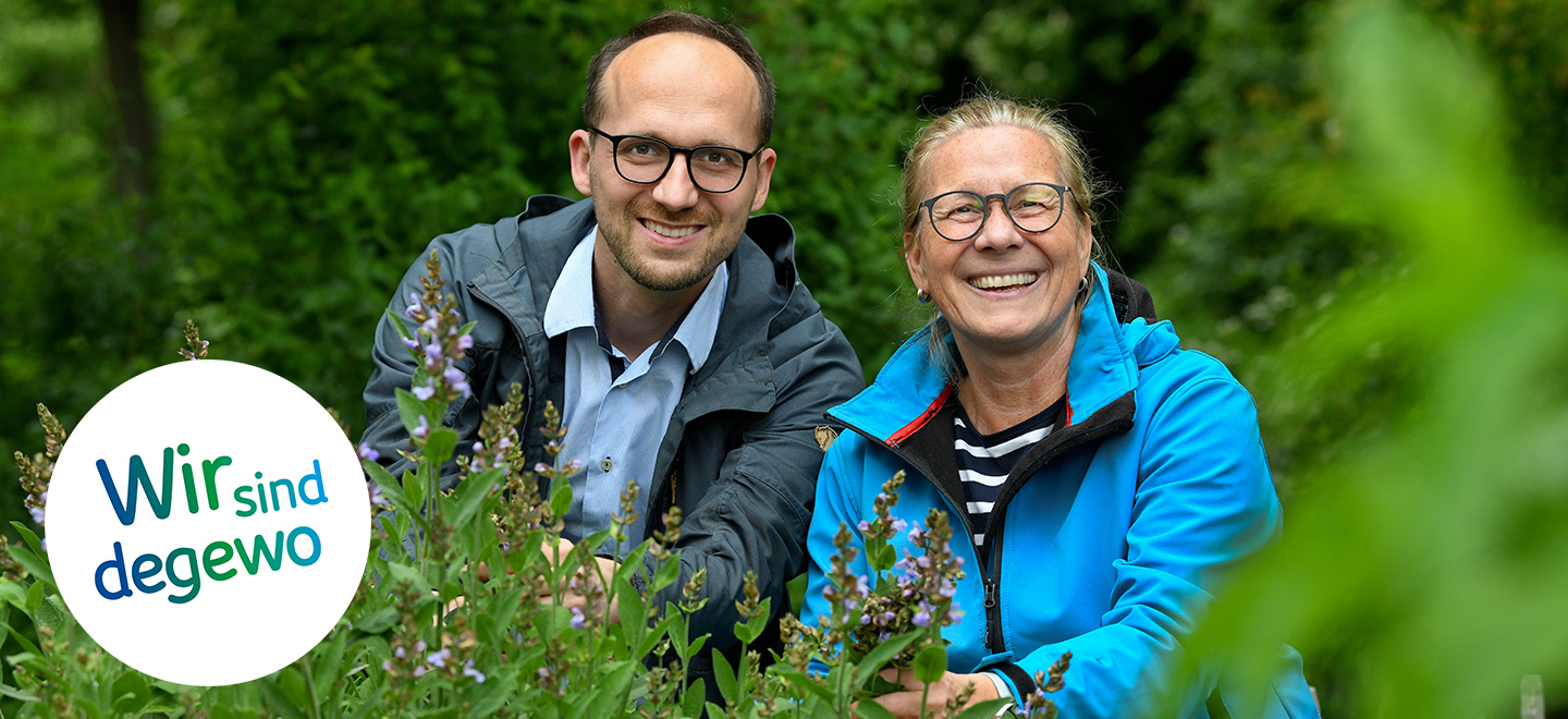 Zwei Mieter sitzen im hohen Gras ihres Gemeinschaftsgartens und lächeln in die Kamera.
