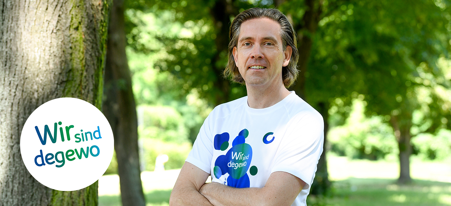 degewo Mitarbeiter Christopher Schneider steht in einem Park neben einem Baum. Er trägt ein Sport T-Shirt, seine Arme sind verschränkt. 