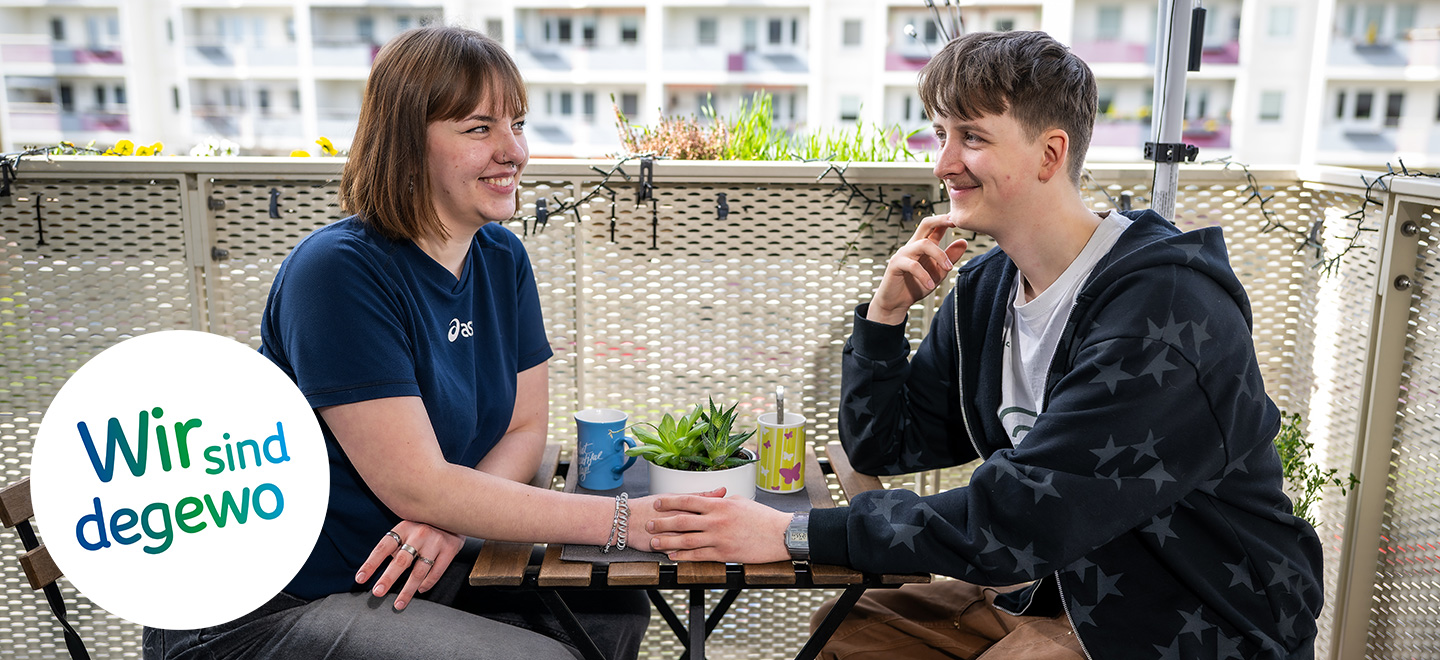 Ein junges Paar sitzt auf dem Balkon auf Stühlen gemeinsam an einem Tisch. Sie schauen sich in die Augen und halten Händchen. Im Hintergrund sind verschwommen Gebäude im Plattenbaustil zu erkennen. Es ist Frühling, einige Pflanzen sind auf dem Balkon gepflanzt.