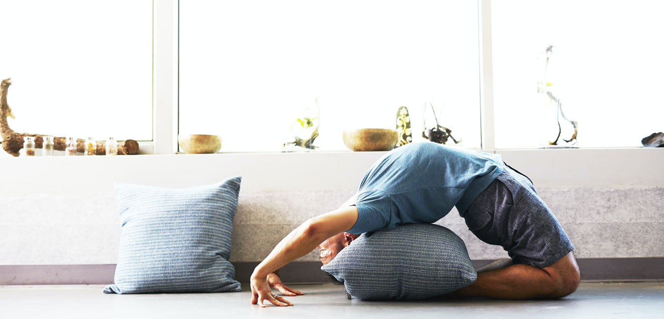 Ein Mann praktiziert Yoga in einer Backbend-Pose. er hat ein graues Kissen, um auf seinem Rücken zu ruhen. Hinter ihm befindet sich ein Fensterbrett, das mit einer Reihe von Schalen und Pflanzen geschmückt ist. Auf dem Boden liegt auch ein Kissen in der gleichen Farbe.