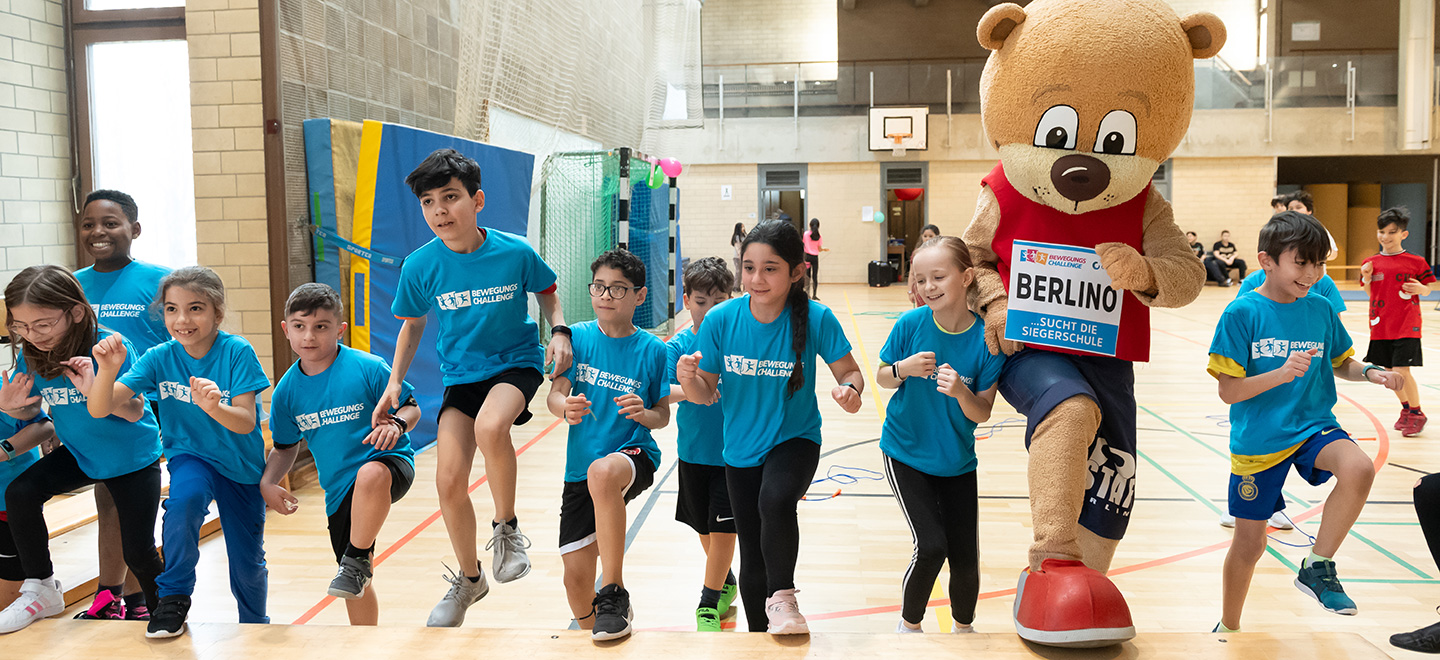 Das Maskottchen Berlino der degewo-Bewegungschallenge rennt mit Kindern einer Grundschule durch die Sporthalle. Sie halten sich an den Händen. 