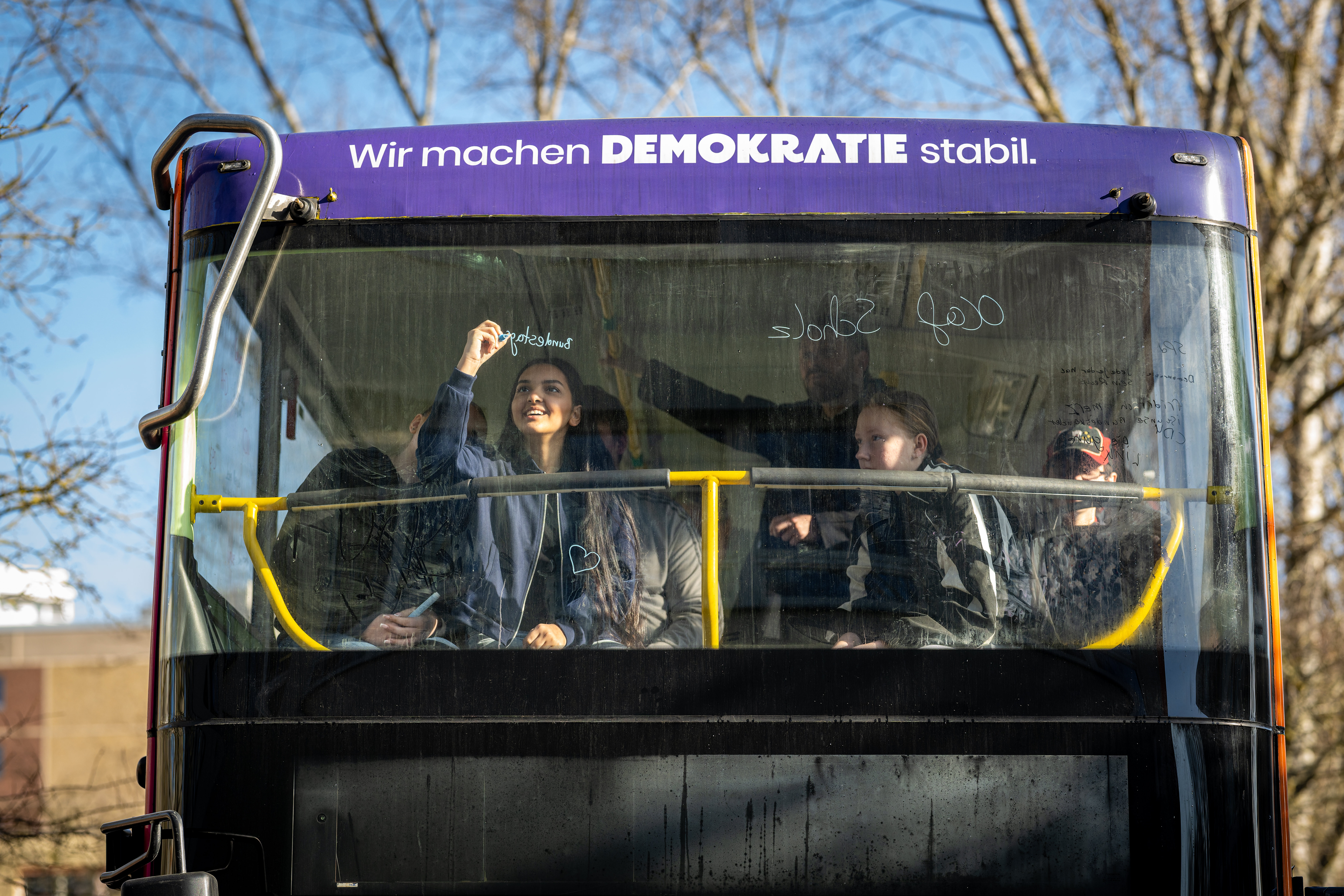 Blick von Außen auf die Frontscheibe eines Doppeldecker-Busses mit der Aufschrift: Wir machen Demokratie stabil. Hinter der Scheibe schreibt ein Mädchen etwas ans Fenster mit einem bunten Stift.
