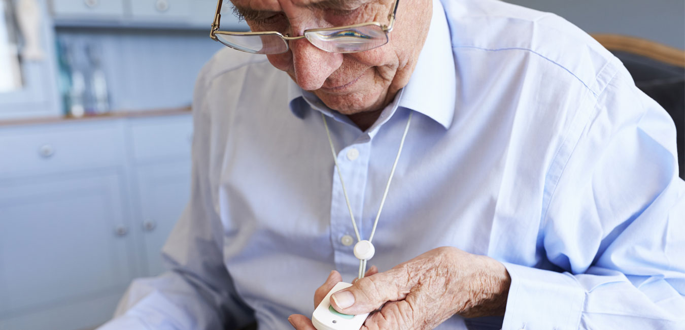 Ein älterer Herr schaut auf sein Band mit dem Notrufknopf, welches um seinen Hals hängt.