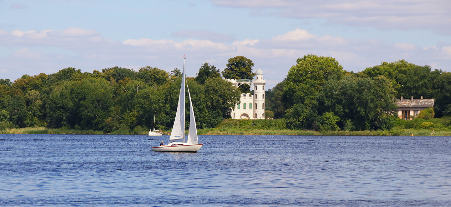 Zu sehen ist der Berliner Fluss Havel im Sommer. Mehrere Segelboote sind auf dem Wasser unterwegs. Im Hintergrund ist die Pfaueninsel und das darauf befindliche Schloss zu sehen. 