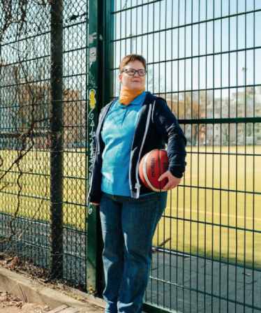 Eine leger gekleidete Frau mit Basketball unter dem Arm lehnt am Zaun eines Sportplatzes.