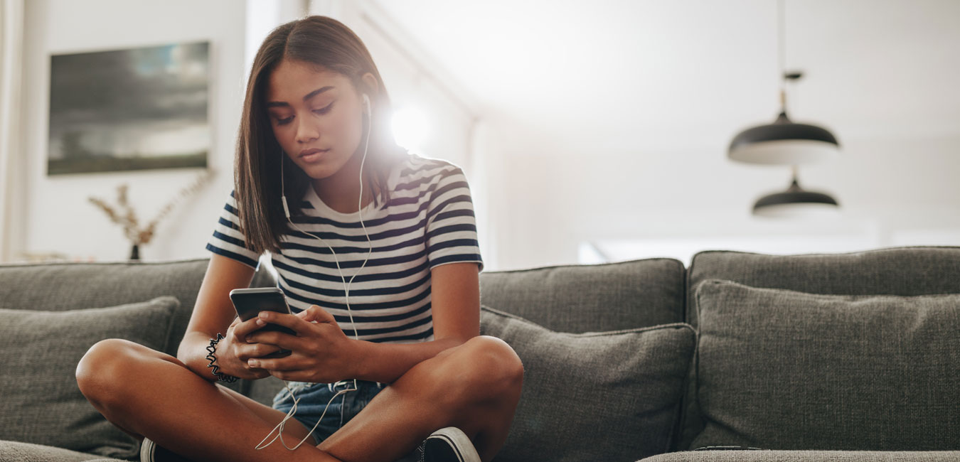 Ein Mädchen in schwarz-weiß Freizeitkleidung sitzt auf dem grauen Sofa und hört Musik über kabelgebundene In-Ear-Kopfhörer