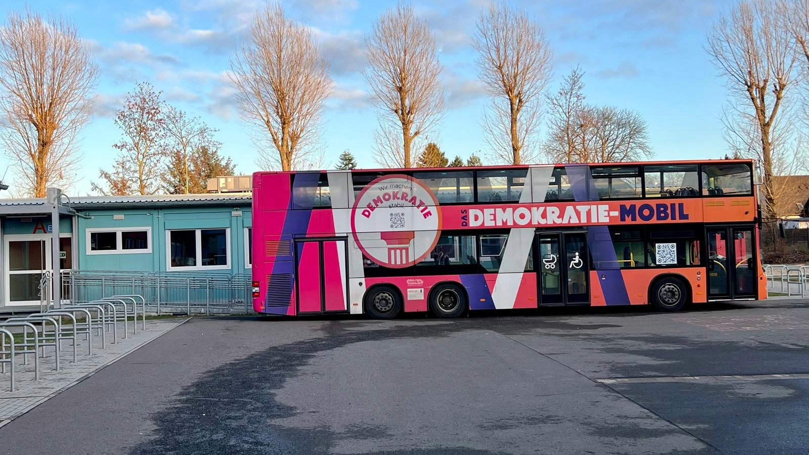 Bunt gestalteter Doppeldeckerbus "Demokratie-Mobil" steht vor Gebäuden und Bäumen.