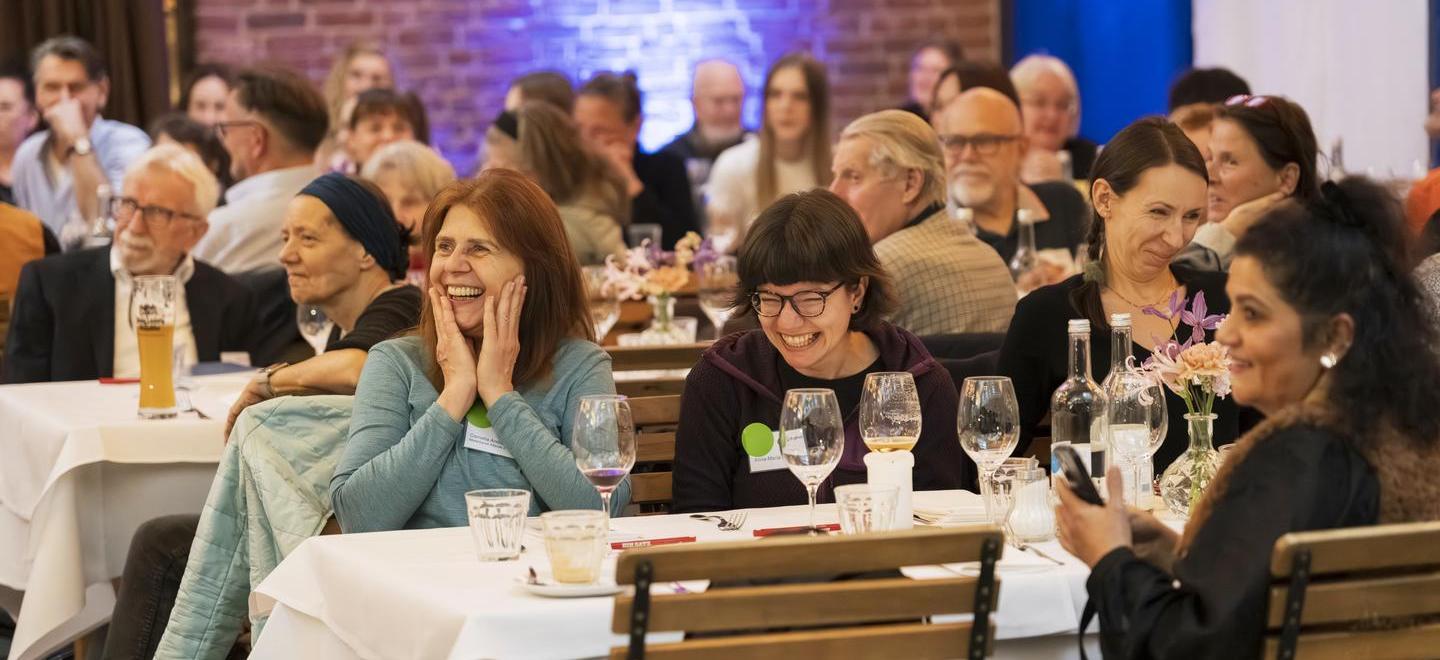 Teilnehmende Mietergremien sitzen am Tisch und lachen herzhaft. Sie schauen dem Bühnenprogramm zu.