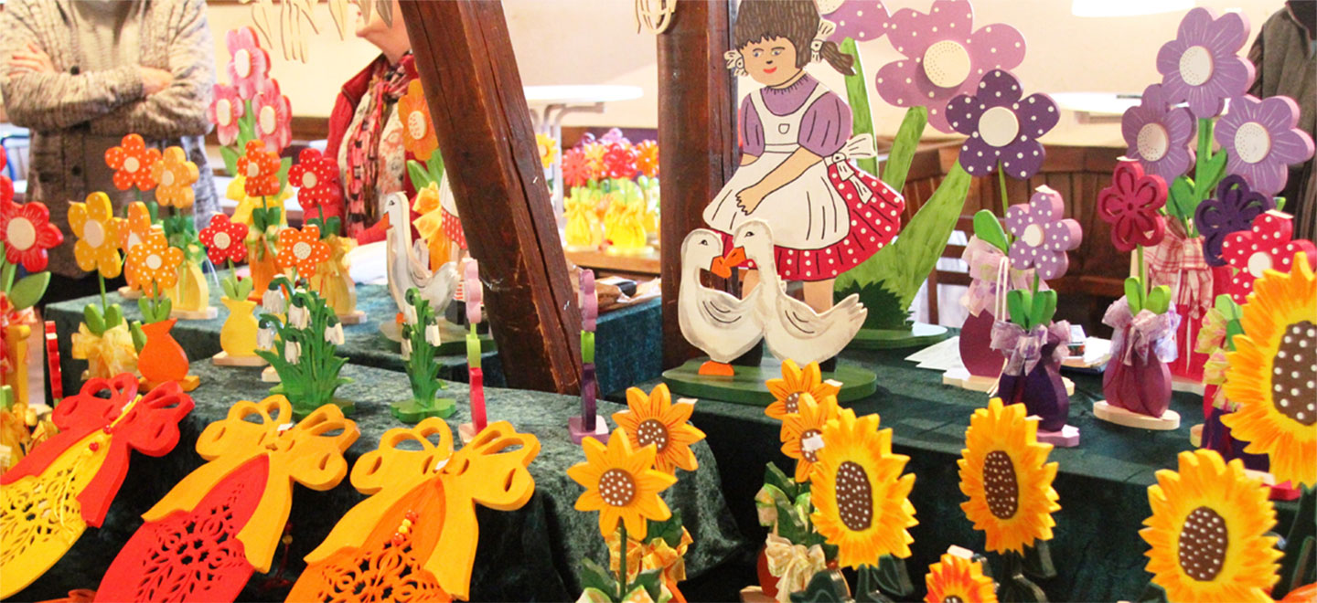 Blick in einen Marktstand mit farbenfroher Osterdekoration wie Hasen und Blumen. Im Hintergrund sind verschwommen die Marktbetreiber zu sehen.