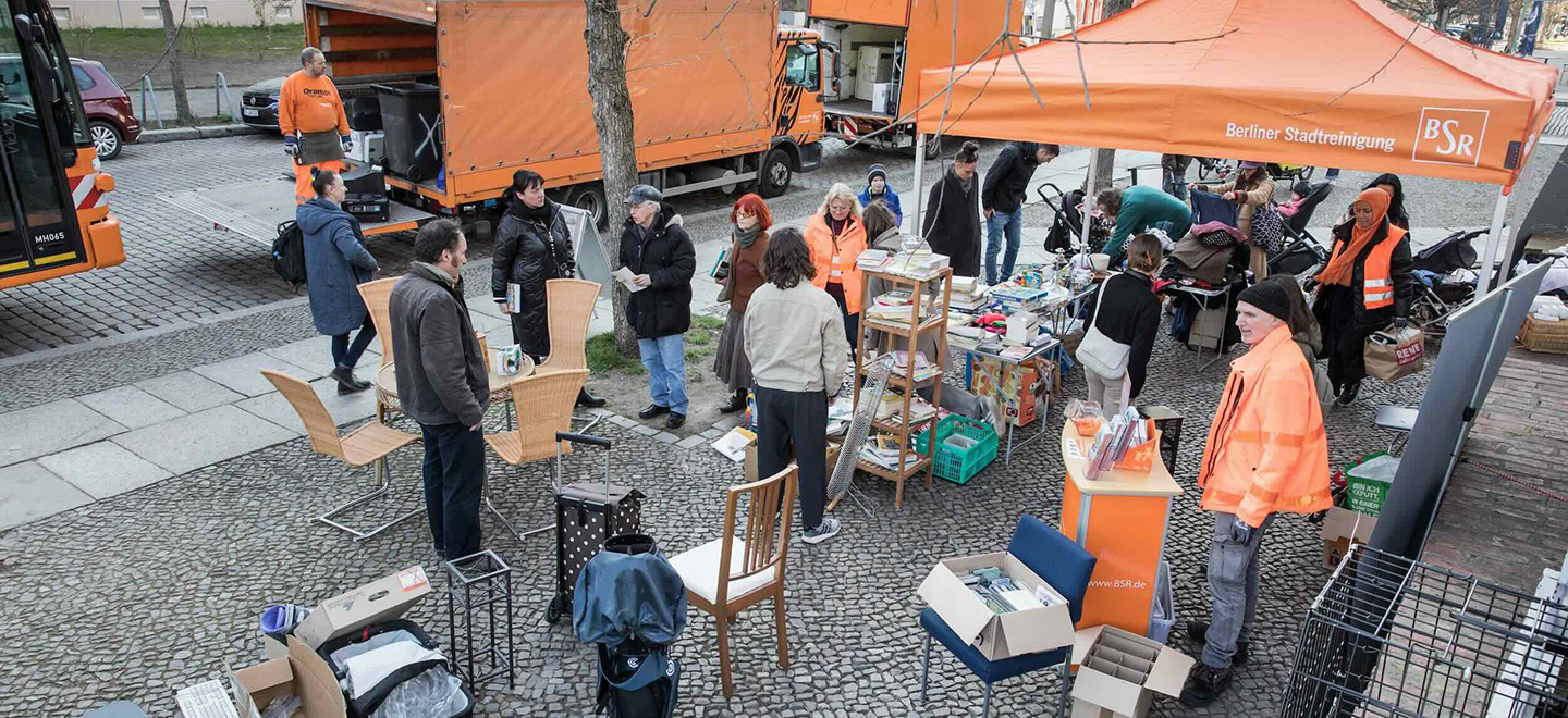 Das Bild zeigt eine Szene von einem BSR-Kieztag. Im Vordergrund stehen diverse Möbelstücke und Haushaltsgegenstände auf einem gepflasterten Platz. Mehrere Personen durchstöbern die Gegenstände oder unterhalten sich. Unter einem orangefarbenen Pavillon mit der Aufschrift “Berliner Stadtreinigung" und dem Logo der BSR sind weitere Gegenstände ausgestellt. Im Hintergrund sind orangefarbene BSR-Lastwagen und Mitarbeiter der BSR in orangefarbener Arbeitskleidung zu sehen. Die Atmosphäre wirkt geschäftig und gemeinschaftlich, während die Teilnehmenden die Gelegenheit nutzen, sich von nicht mehr benötigten Gegenständen zu trennen und diese weiterzuverwenden.