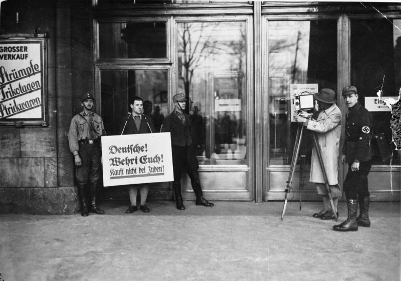 Boykottaktion der Nazis gegen jüdische Geschäfte, Filmleute warten auf Publikum, welches das Warenhaus Wertheim betreten will. SA- und SS-Leute stehen davor, ein Mann mit Schild um den Hals „Deutsche! Wehrt Euch! Kauft nicht bei Juden!“, rechts Mann mit Filmkamera filmend.