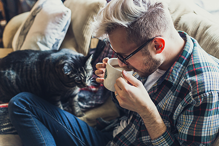 Ein Mann trinkt Kaffee auf seinem Sofa, daneben gibt es eine schwarze Katze