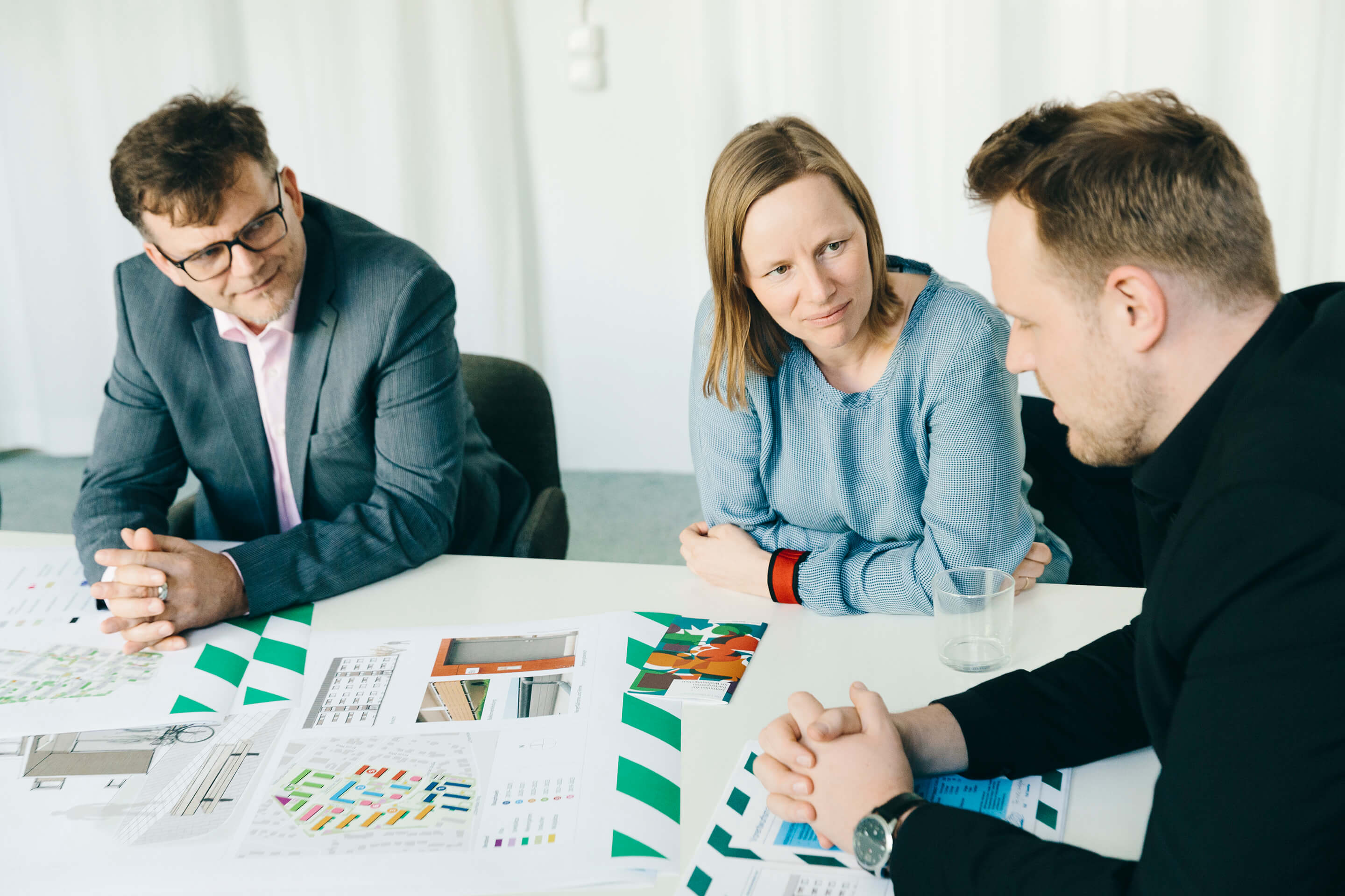 Drei Kollegen sitzen am Tisch und diskutieren mit Plänen vor sich liegend.