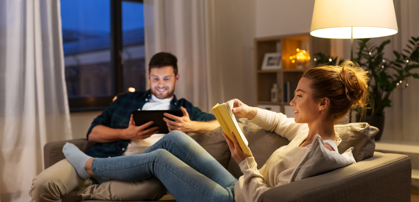 Eine Frau und ein Mann sitzen auf der Couch. Der Mann schaut auf sein Tablet und die Frau liest ein Buch. Im HIntergrund steht eine Lampe.