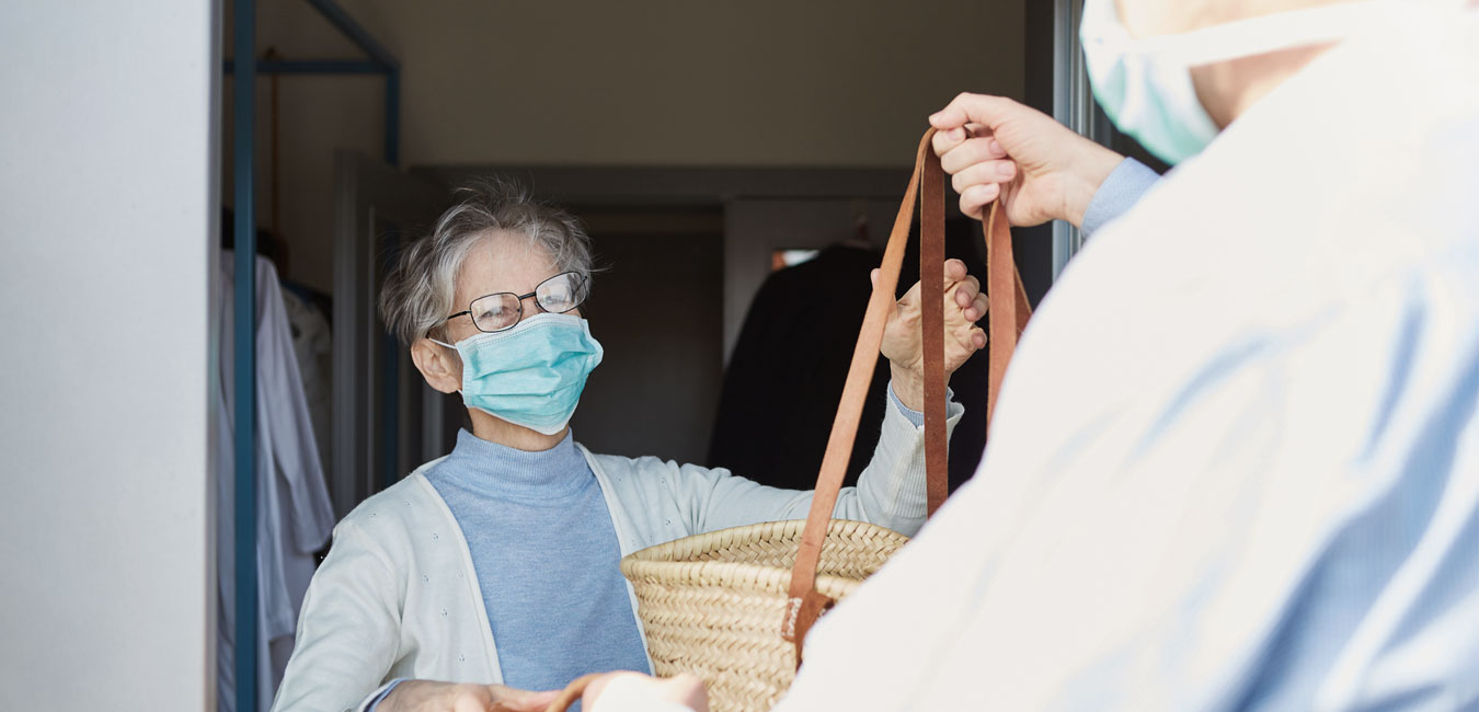 Das Bild zeigt, wie ein Mann einer älteren Frau Einkäufe in Tragekörben übergibt. Beide tragen zum Schutz vor Corona einen Mund-Nase-Schutz.