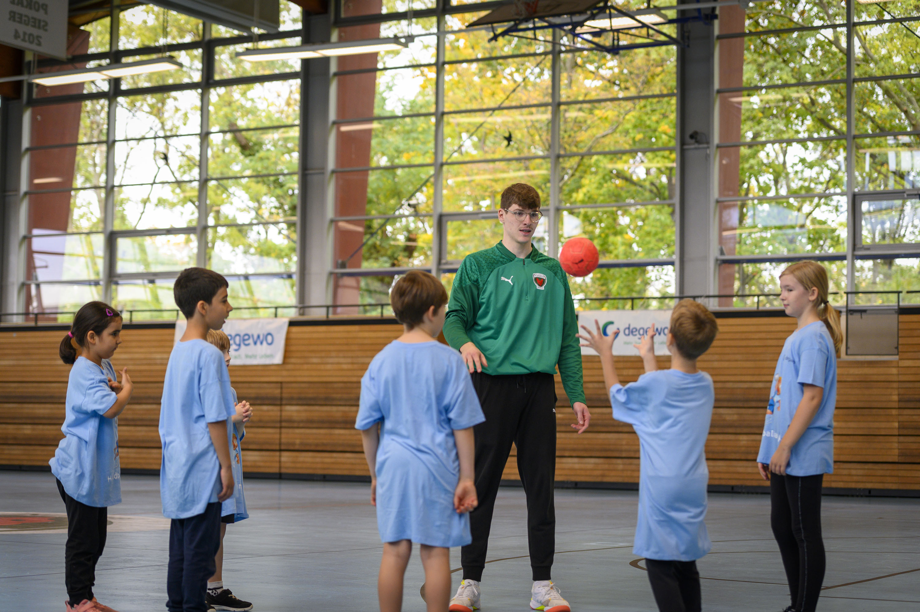 Eine Gruppe Kinder steht in einer Sporthalle mit ihrem Trainer. Ein Kind wirft einen kleinen roten Ball in Richtung Trainer.